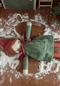 Fabric doll with a green dress, red hood, and soft facial features, lying on a wooden surface dusted with flour, creating a snow angel shape.