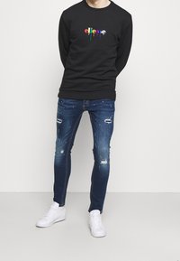 Felpa nera con logo multicolore, jeans in denim blu slavato e strappato, e sneakers bianche, che mostrano una vestibilità slim e uno stile casual.