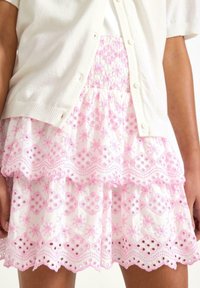Cardigan bianco sopra una gonna a balze ricamata rosa, con motivi floreali e intagli ad occhio lungo l'orlo. Tessuto morbido.