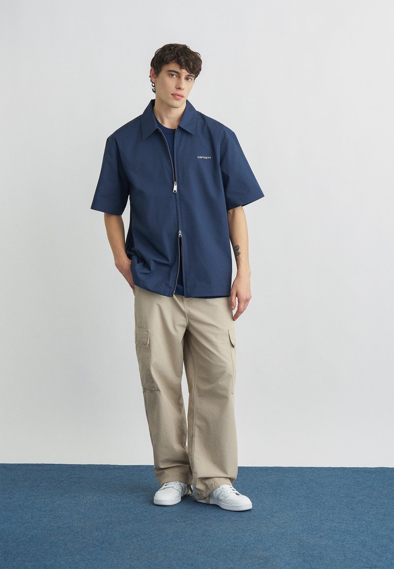 Jeune homme aux cheveux bouclés portant une chemise zippée à manches courtes bleu marine, un pantalon cargo beige et des baskets blanches, debout sur un tapis bleu.