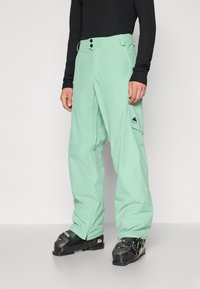 Pantaloni da neve verde menta con una vestibilità ampia, realizzati in materiale impermeabile. Presentano due bottoni frontali, tasche laterali e un piccolo logo sulla gamba.