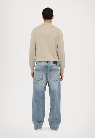 Μπεζ πουκάμισο με μακριά μανίκια, συνδυασμένο με χαλαρά, ανοιχτό μπλε τζιν παντελόνια που διαθέτουν δύο πίσω τσέπες και μια μαύρη ετικέτα μάρκας στη μέση.