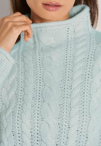Frau trägt einen hellblauen, grob gestrickten Rollkragenpullover und hält mit der linken Hand den Kragen fest, Lippen sind oberhalb des Ausschnitts sichtbar.