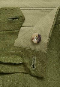 Olivově zelená látka s texturou rybí kosti, s knoflíkovým zapínáním a vyšitými detaily. Viditelná kapsa na zip.