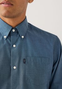 Chemise boutonnée en tissu bleu foncé, avec un col pointu, des boutons blancs, une poche plaquée et une petite étiquette.
