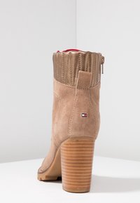 Zamszowy botek za kostkę w jasnobrązowym kolorze, z metalicznym złotym akcentem na górze, prążkowaną fakturą oraz piętą z drewnianych warstw. Posiada boczny zamek błyskawiczny.