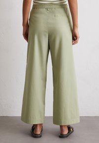 Persoon in lichtgroene wijde broek en zwarte sandalen, staande op een grijze vloer tegen een witte gestructureerde muur.