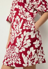 Robe florale rouge avec imprimé botanique blanc, manches courtes, poches latérales, et coupe décontractée avec fentes en bas. Fabriquée en tissu léger.