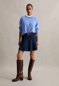 Mulher com cabelo trançado, vestindo um suéter azul claro, uma saia azul escura com cinto castanho e botas de couro castanhas altas, de pé contra um fundo simples.