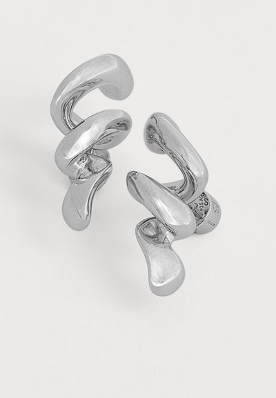 Paire de boucles d'oreilles puces brillantes en argent de forme abstraite avec des surfaces lisses et courbes sur un fond clair uni.