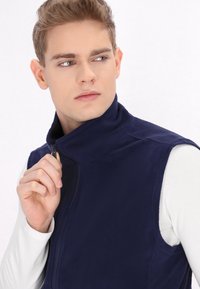 Mladý muž v námorníckej modrej fleecovej veste na bielom dlhom rukáve, zapínajúci vestu, pozerajúci sa na stranu na bielom pozadí.