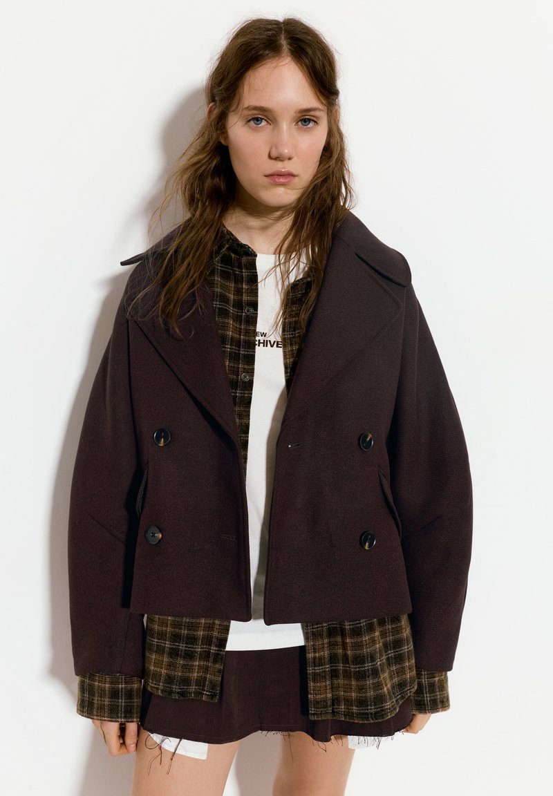 Manteau cropped double boutonné marron foncé avec de grands revers, porté sur une chemise à carreaux et un T-shirt blanc, avec des bords effilochés à l'ourlet.