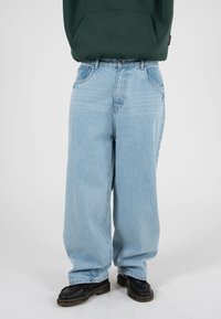 Hele sinised laia lõikega teksapüksid, millel on lõõgastunud istuvus, kõrge vöökoht, viis taskut ja toonitooniline nööp. Kandmisel mustad jalanõud.