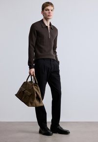 Rudas megztas megztinis su užtrauktuku ant apykaklės, juodos kelnės ir juodi batai. Modelis laiko rudą zomšą rankinę su priekinėmis kišenėmis ir užtrauktukais.