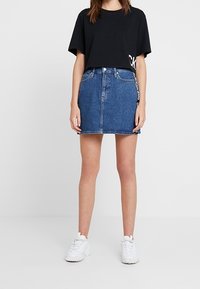 Kvinna som bär en svart kortärmad croppad t-shirt, blå denim mini kjol med framfickor och vita sneakers, stående på en enhetlig bakgrund.