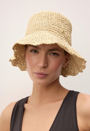 Femeie purtând o pălărie tip borsetă bej, țesută, și un top negru fără mâneci, privind înainte cu o expresie neutră pe un fundal deschis.