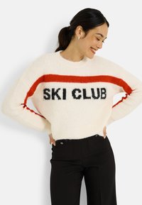 Suéter blanco y peludo con una franja roja y la inscripción negra "SKI CLUB" en el pecho. Combinado con pantalones negros de talle alto.