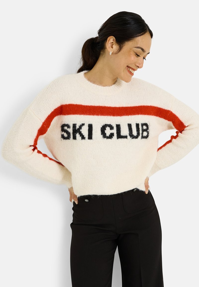 Suéter blanco y peludo con una franja roja y la inscripción negra "SKI CLUB" en el pecho. Combinado con pantalones negros de talle alto.