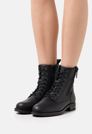 Svart läderkängor i kamplängd, med snörning och sidogdetaljdragkedja, bärs på bara ben mot en enkel vit bakgrund.