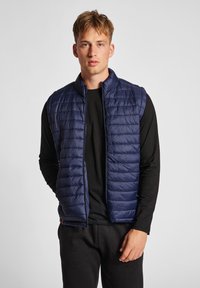 Marinblå pufferväst med horisontell quiltad textur, hög krage och frontdragkedja. Bärs över en svart långärmad tröja.