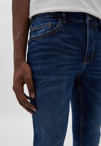 Nära bild av en persons hand som vilar bredvid blå denimjeans med orange sömmar och en synlig framficka.