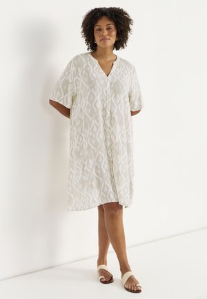 Kvinde med krøllet hår iført en beige knælang kjole med mønster og knapper samt beige åbne sandaler, står mod en hvid baggrund.