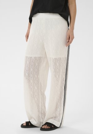 Białe szerokie spodnie szydełkowane z czarnymi paskami po bokach, noszone z czarnymi sandałami i czarnym topem na stojącej osobie.