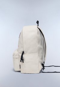 Béžový batoh vyrobený z odolné tkaniny s černými popruhy, boční kapsou a stahovacími uzávěry. Na přední straně má decentní logo.