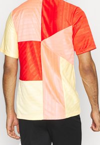 Kortärmad t-shirt med ett lapptäcksdesign i röd, orange, persika och gul färg med en glänsande textur och subtila varumärkesmönster.