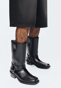 Person wearing black mid-calf leather boots with square toes and straps, paired with knee-length black shorts on a white background.