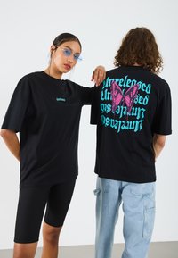 Black oversized t-shirts feature a pink butterfly graphic and blue text on the back. The model on the left wears black bike shorts.