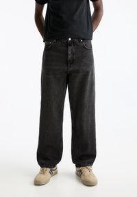 Jeans in denim nero con una vestibilità ampia, caratterizzati da gamba dritta e due tasche frontali. Abbinati a sneakers beige e a una maglietta nera.