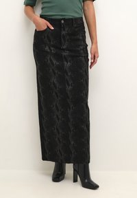 Jupe longue en denim noir avec un motif serpent, poches avant, taille cintrée et fermeture à bouton ; associée à des talons noirs.