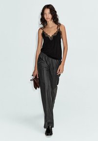 Schwarzes Top mit Spitze kombiniert mit grauen Hosen im großzügigen Streifenmuster. Das Model hält eine braune Tasche; die Schuhe sind schwarz und glänzend.