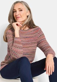 Maglione lavorato a maglia con un motivo multicolore in rosa, verde e crema. Collo rotondo e polsini a coste. Modello abbinato a pantaloni blu scuro.