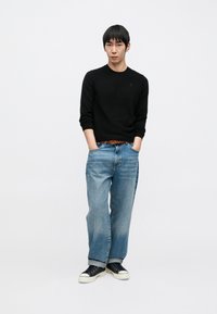 Juodas ilgarankovė megztinis, šviesiai mėlynos platėjančios kelnės, rudas diržas, juodi sportbačiai su baltais padais. Vienodas fonas pabrėžia produkto išskirtinumą.