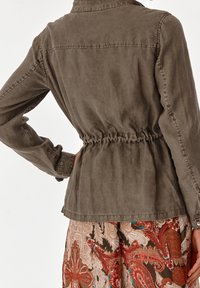 Person som bär en brun jacka med insvängd midja över en flerfärgad paisleymönstrad kjol, sedd bakifrån med ena handen på höften.