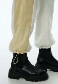 Schwarze Lederankelstiefel mit dicken, strukturierten Sohlen, kombiniert mit cremefarbenen Hosen, die eine glatte, glänzende Seitenpartie und geraffte Bündchen aufweisen.