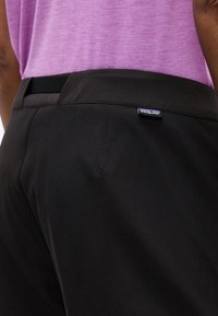 Κοντινό πλάνο ατόμου που φοράει μαύρο παντελόνι Patagonia και μοβ πουκάμισο, εστιάζοντας στην περιοχή της μέσης με εμφανή την ετικέτα της μάρκας.