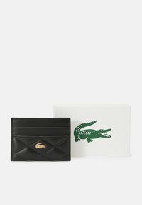 Portafoglio in pelle nera trapuntata con logo di coccodrillo dorato, posizionato davanti a una scatola bianca con un emblema di coccodrillo verde.