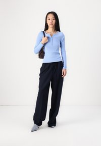 Lyseblå langermet henley-skjorte, marineblå vide bukser og grå pumps. Modellen bærer en liten brun håndveske. Ensfarget bakgrunn.