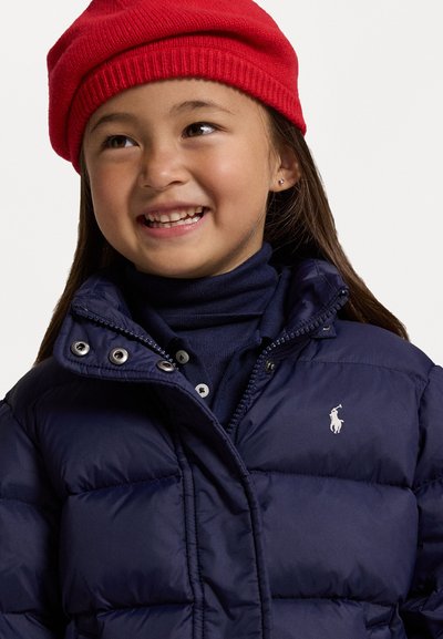 Navy puffer jacket with quilted texture and silver hardware, layered over a navy turtleneck; paired with a red knit beanie.