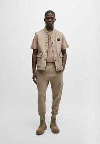 Beige ärmellose Weste über einem hellbraunen Hemd, kombiniert mit beige farbenen Cargo-Hosen und braunen Schnürstiefeln. Die Weste hat zwei Vordertaschen.