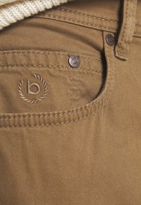 Bruine katoenen broek met een gestructureerde afwerking, voorzien van een klein geborduurd logo, metalen knoopdetails en zichtbare stiksels langs de randen.