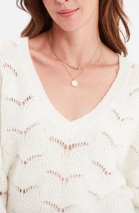 Maglione bianco lavorato a maglia con design a onde e scollatura a V profonda. Abbinato a due collane d'oro a catena, una delle quali con un pendente rotondo.