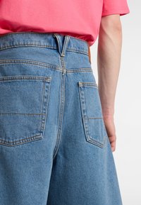 Jasnoniebieskie krótkie szorty dżinsowe z dwoma tylnymi kieszeniami, widocznymi przeszyciami i brązową skórzaną metką na pasku.