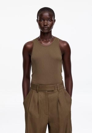 Kobieta o ciemnej karnacji, ubrana w oliwkowozielony, bezrękawnik i plisowane spodnie z wysokim stanem, stojąca z rękami w kieszeniach na białym tle.