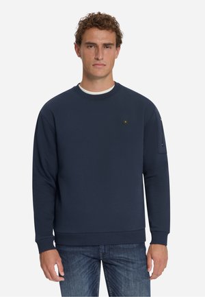 Man met krullend haar die een donkerblauwe lange mouwen trui en een blauwe spijkerbroek draagt, staand tegen een effen lichte achtergrond.