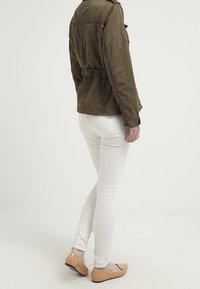 Giacca belted verde oliva con una leggera lucentezza, abbinata a jeans skinny bianchi e ballerine color carne; texture liscia, design aderente.