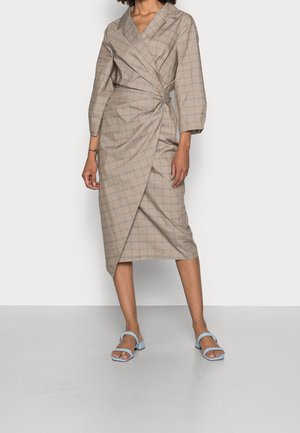 Femme portant une robe portefeuille beige à motif discret à carreaux et manches trois-quarts, associée à des sandales bleu clair ouvertes à l\'orteil.